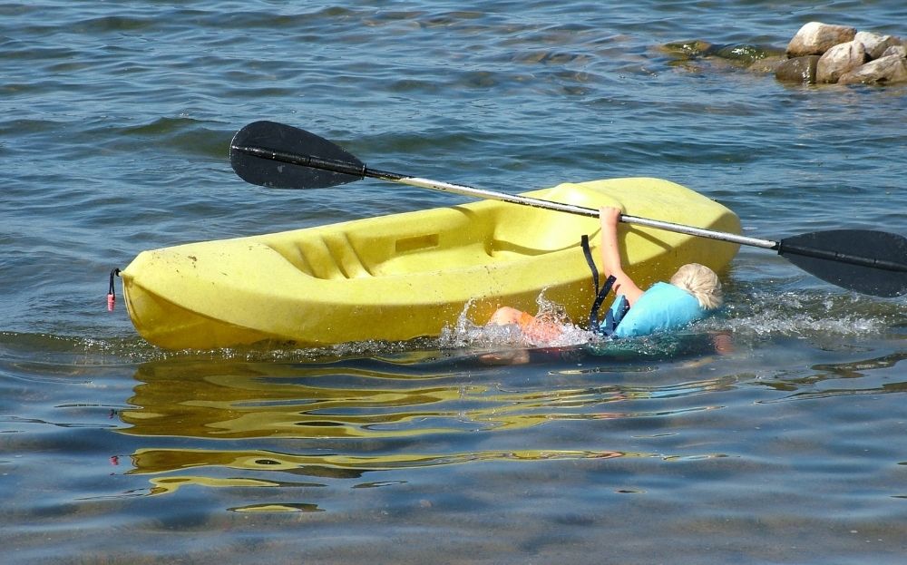 Man falling overboard from a recreational kayak. It is shallow water near the coast and the man is wearing a safety lifejacket.